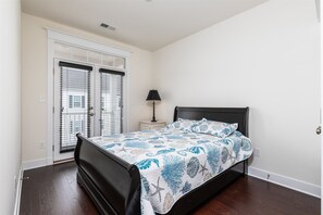 A bedroom with a bed, a nightstand, a lamp, and a window with blinds. - Broad Marsh Unit 112E (Ocean City)