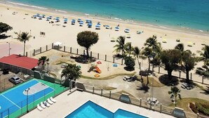 A beach view with a pool area, palm trees, and beach umbrellas.