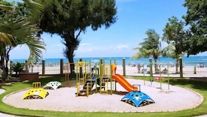 A playground with slides and climbing structures, surrounded by a sandy area and trees, with a beach and ocean in the background.