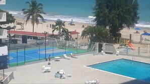 A pool area with a tennis court, lounge chairs, and a beach in the background.