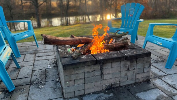 Un foyer avec des bûches qui brûlent, entouré de chaises bleues, installé sur une terrasse en briques avec une vue sur un lac en arrière-plan.