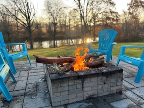 Un foyer avec des bûches qui brûlent, entouré de chaises bleues, installé sur une terrasse en briques avec une vue sur un lac en arrière-plan.
