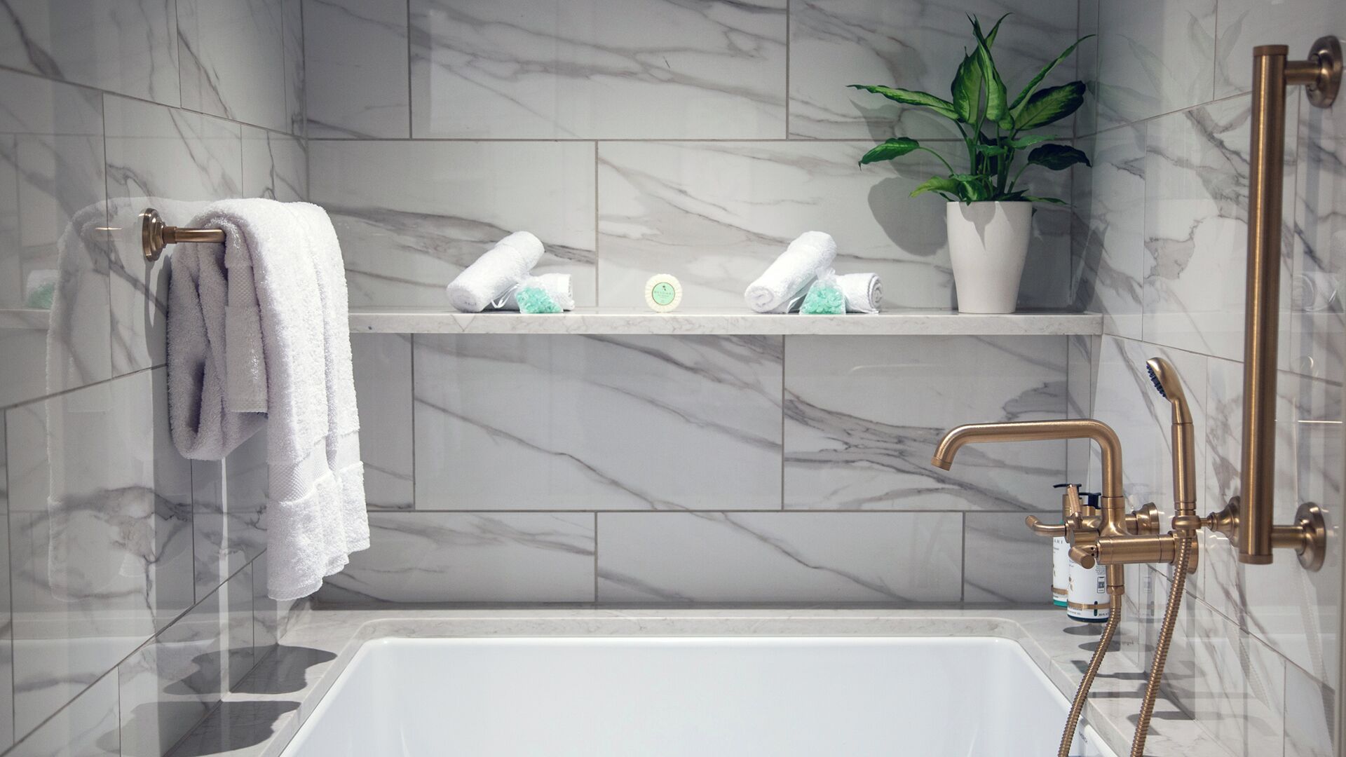 A modern bathroom with a marble wall, a bathtub, a shower with a handheld showerhead, and a shelf with toiletries.