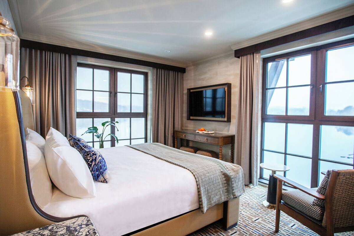 A hotel room with a large bed, a desk, a chair, a TV, and large windows.