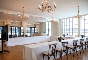 Un amplio salón de banquetes con múltiples mesas dispuestas para comer, elegantes candelabros y grandes ventanales que ofrecen luz natural.