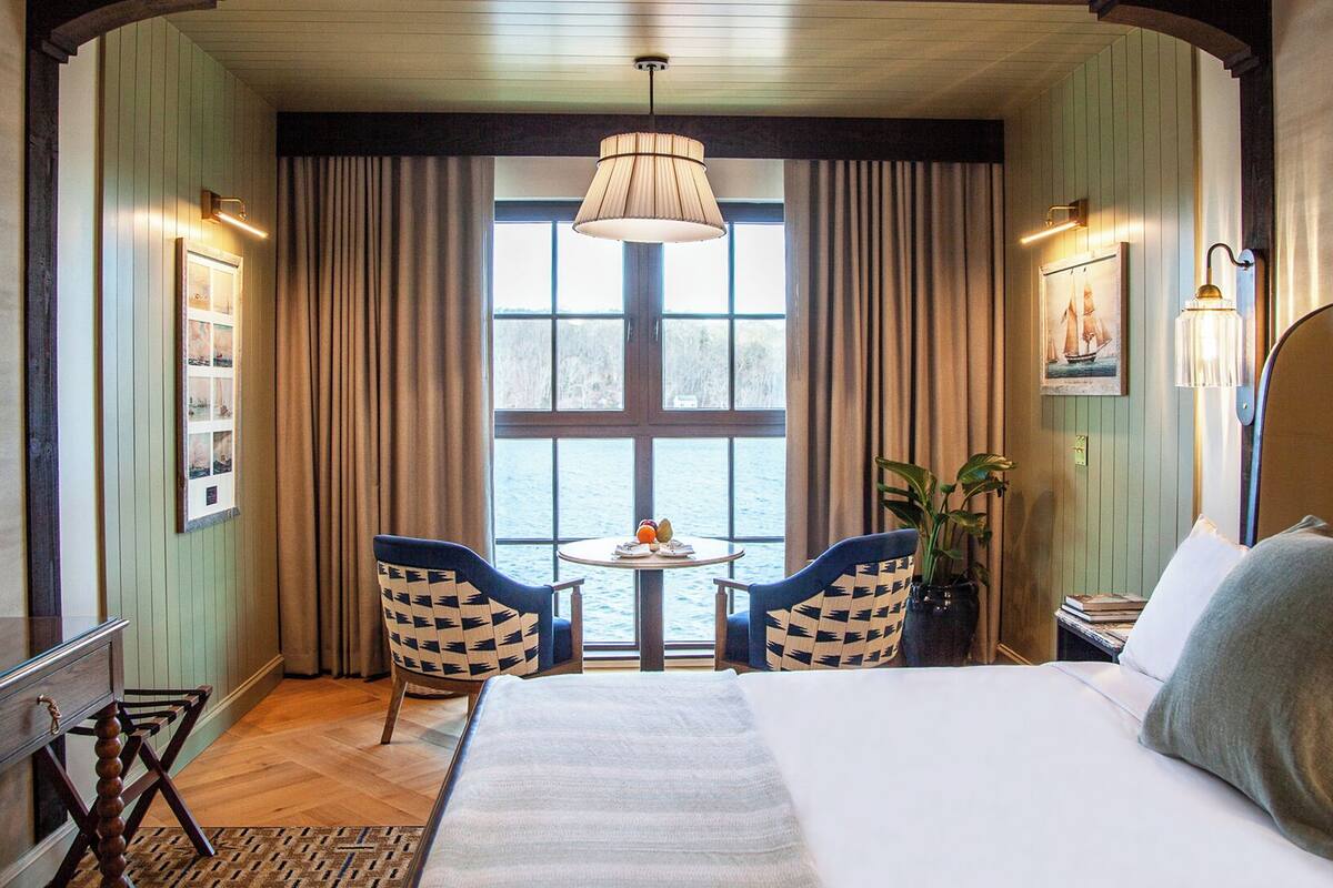 A hotel room with a large bed, two chairs, a small table with fruit, a window with curtains, and a view of the water.