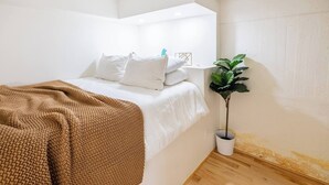 A bedroom with a bed, white bedding, a brown knitted blanket, a potted plant, and a white wall with a small shelf.