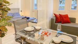 Ein modernes Wohnzimmer mit einem Esstisch aus Glas, gedeckt für zwei Personen, einer Couch mit roten Kissen und einer großen Pflanze.