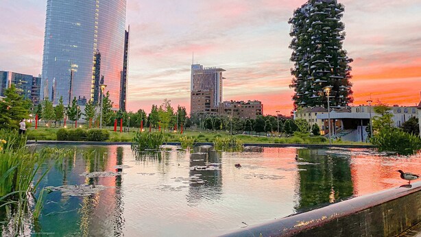 Eine moderne Stadtlandschaft mit einem markanten UniCredit-Gebäude, einem außergewöhnlichen Wohnturm und einem ruhigen Wasserspiel, das den Himmel spiegelt.