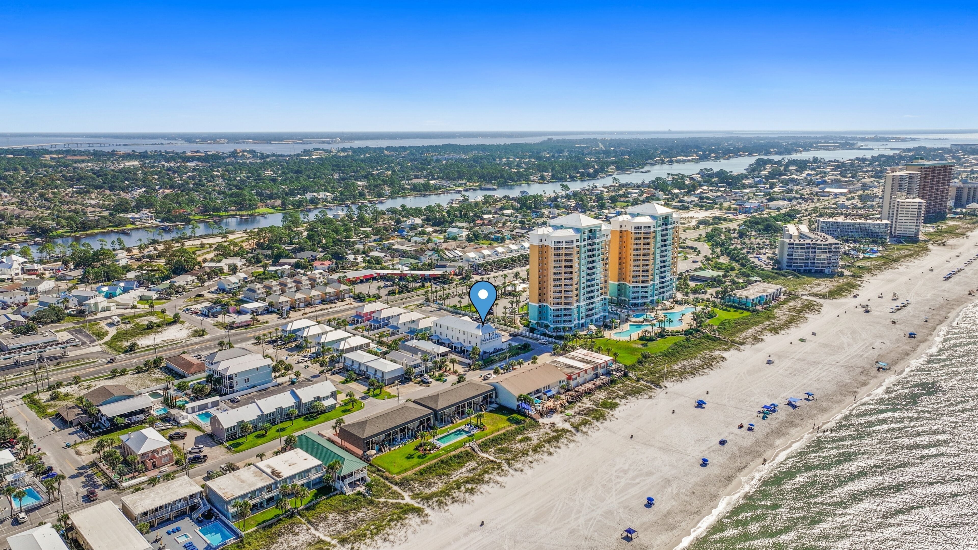 Vista aérea de una ciudad costera con un hotel frente a la playa que cuenta con varias construcciones, una piscina y una playa con sombrillas.