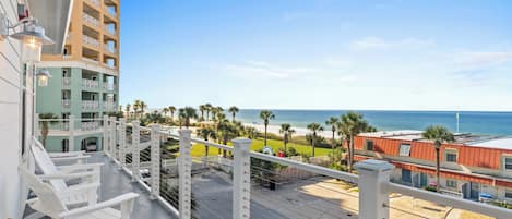 A balcony with white chairs, a view of the beach, and a tall building with balconies.