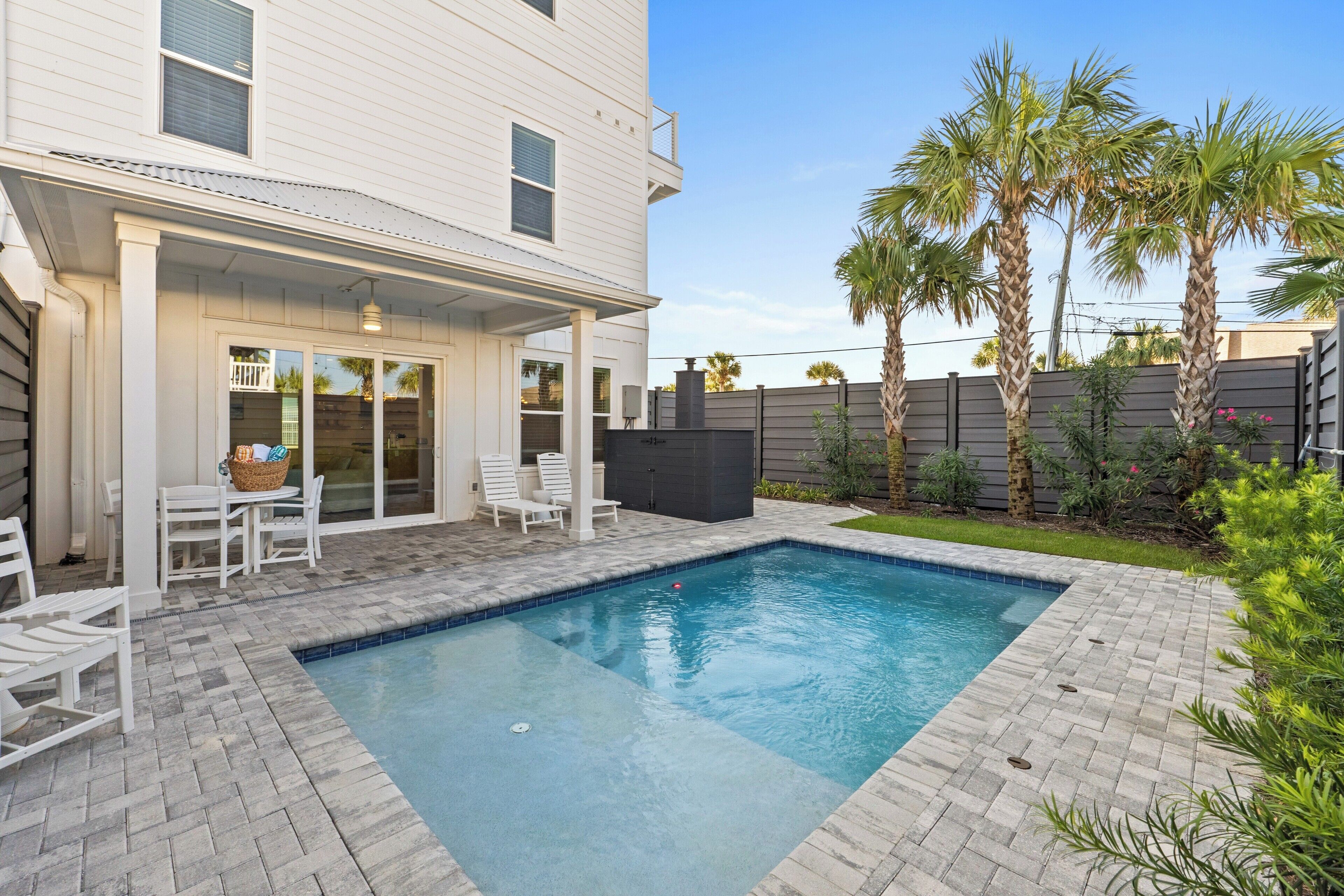 A rectangular swimming pool with a tiled deck, surrounded by a white house with a covered patio and outdoor furniture.
