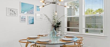 Et spiseområde med et rundt glasbord, seks stole, en vase med blomster og billeder i rammer på væggene.