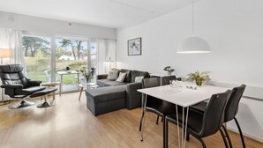 A modern living room with a sofa, a dining table, and a chair. There is a window with curtains and a view of the outdoors.