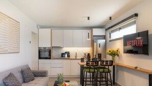 A modern kitchen with a built-in oven, microwave, and coffee maker. There is a dining area with a wooden table and chairs, and a TV mounted on the wall displaying "NETFLIX".