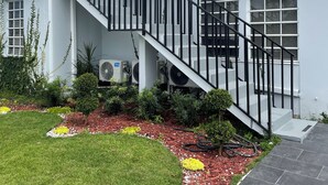Un edificio moderno con fachada blanca, una escalera con barandales negros y un jardín con césped y flores amarillas.