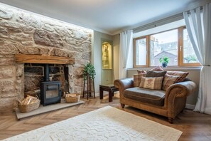 A cozy living room with a stone fireplace, a leather sofa, and a large window with curtains. - Beautifully presented cottage by the beach (Nairn)