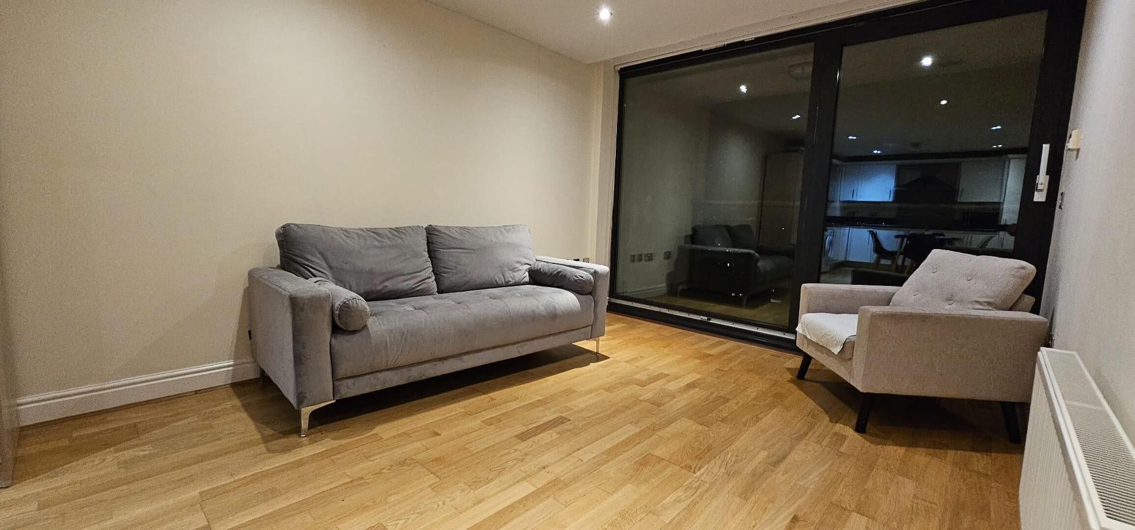 Una sala de estar moderna con un sofá y un sillón grises, suelo de madera y un gran ventanal que da a un balcón.