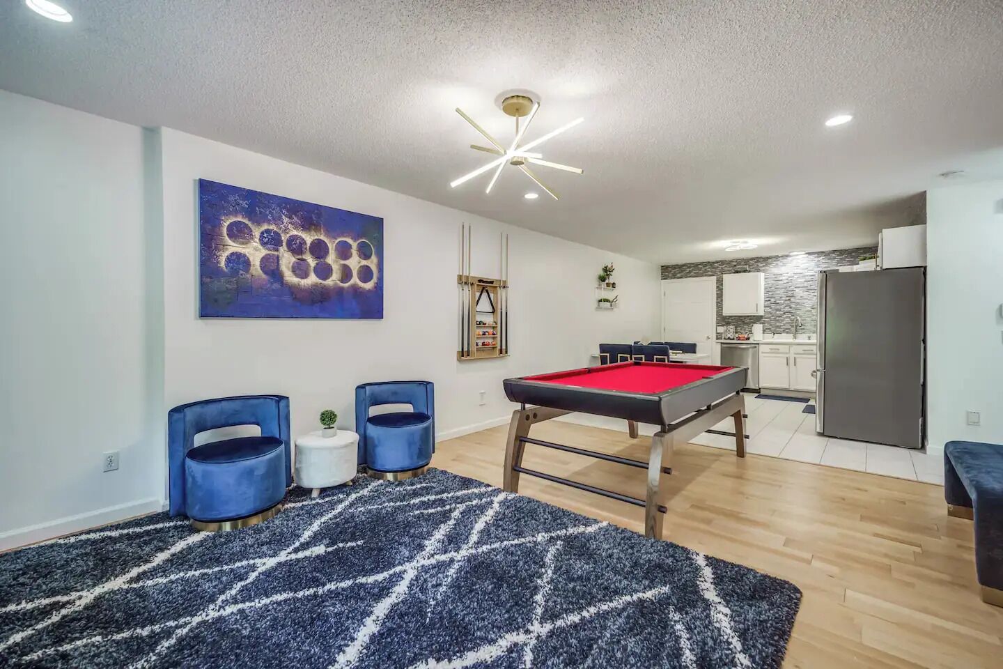 Una sala de estar moderna con mesa de billar, sillas azules, un tapete azul y una cocina al fondo.