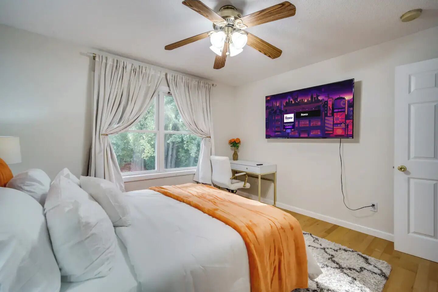 A bedroom with a bed, a desk, a chair, a ceiling fan, a window with curtains, and a wall-mounted TV.