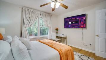 A bedroom with a bed, a desk, a chair, a ceiling fan, a window with curtains, and a wall-mounted TV.