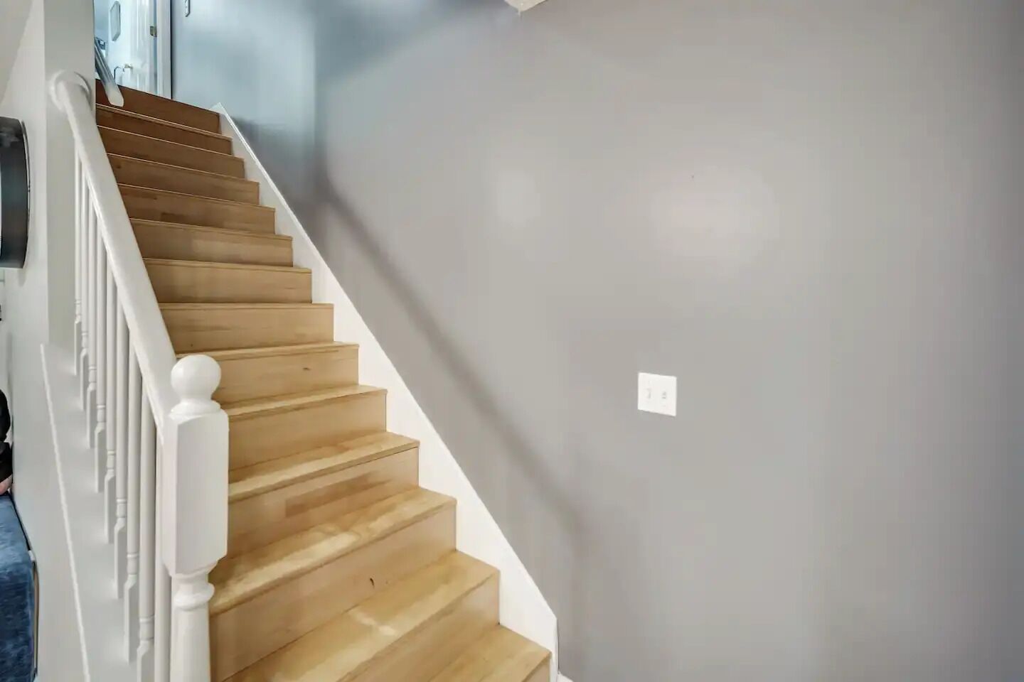 A staircase with wooden steps and white railings, leading to a room with a grey wall and a light switch.