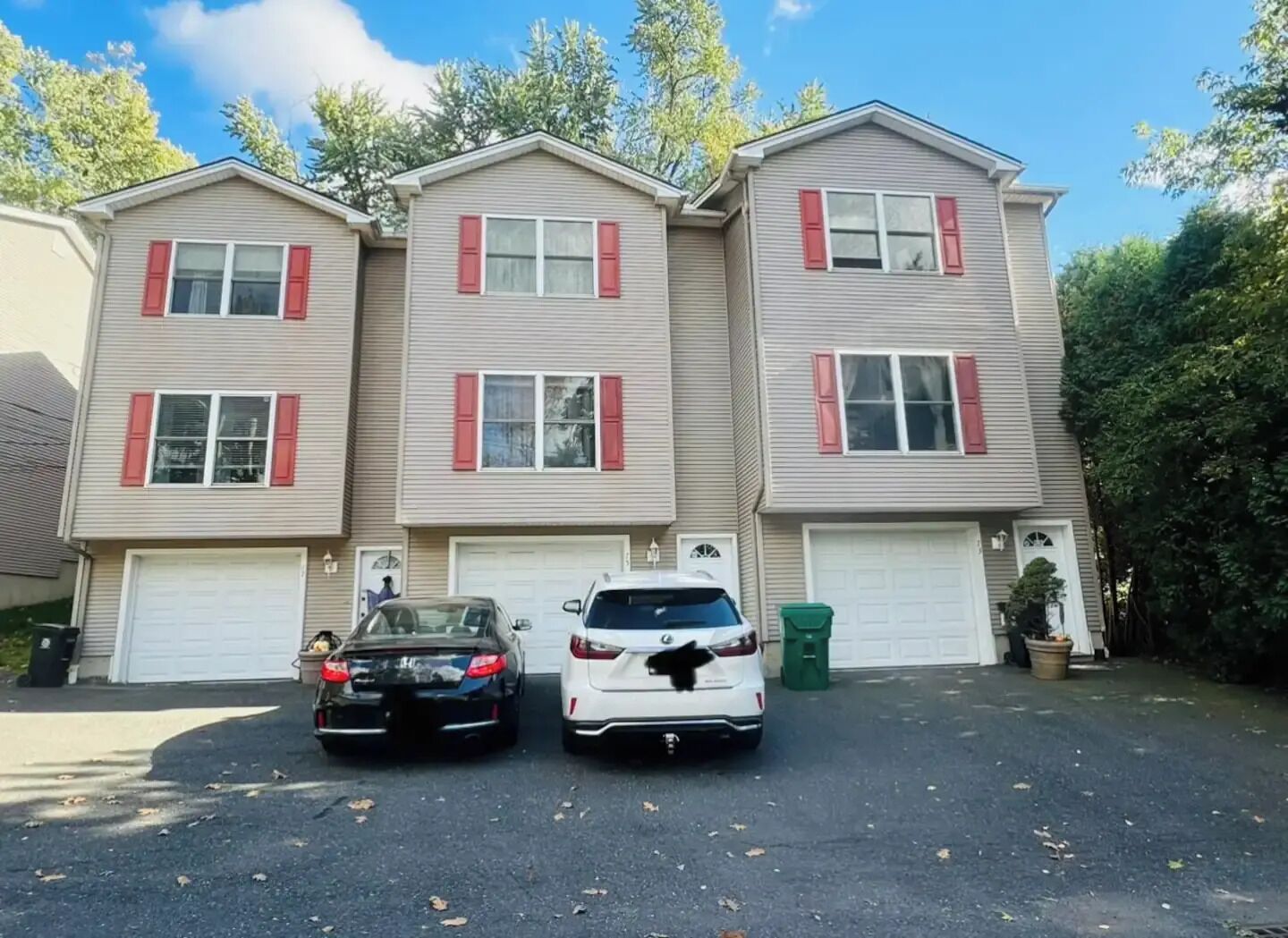 Une maison à deux étages avec un garage, deux voitures garées devant et une poubelle verte.