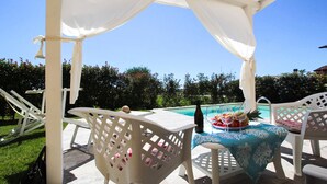 A covered outdoor seating area with a pool, white chairs, and a table with a blue tablecloth.