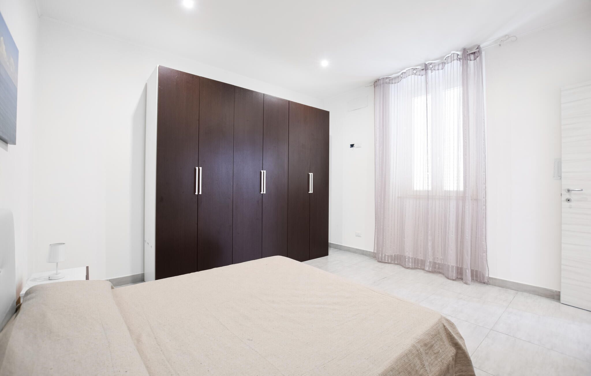 A bedroom with a bed, a wardrobe, a window with sheer curtains, and a door.