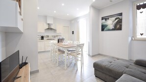 A modern kitchen with white cabinets, a dining area with a wooden table and white chairs, and a gray sofa in a bright living room with a large window and a waterfall painting.