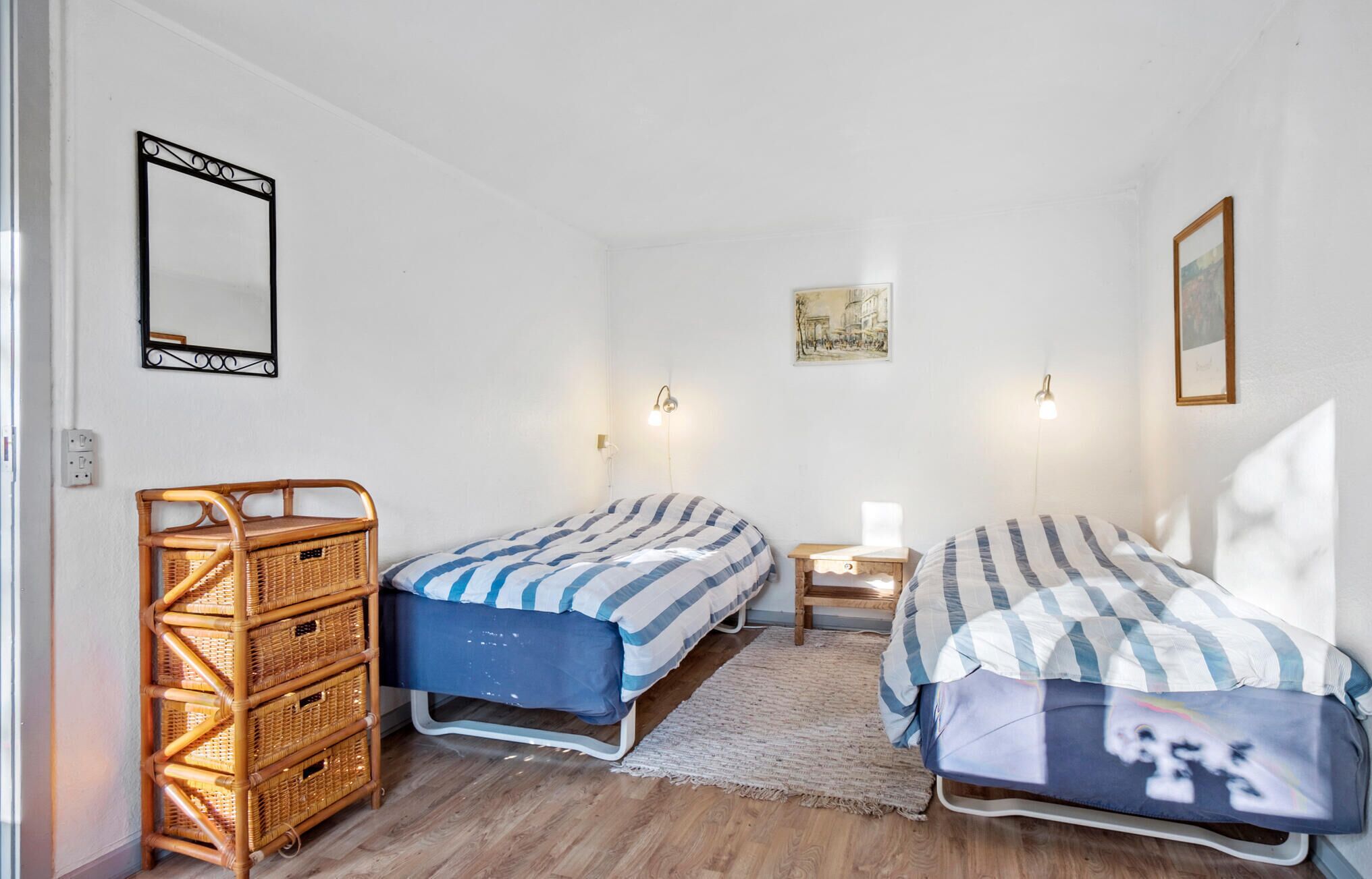A small, well-lit room with two single beds, a wicker chair, a small table, and a wall-mounted mirror.