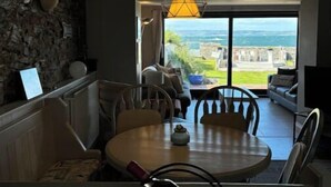 Un coin repas avec une table ronde, des chaises et un luminaire suspendu. Un mur en pierre avec un appareil d’éclairage se trouve sur le côté gauche. Une grande fenêtre donnant sur la mer et les bâtiments se trouve à l’arrière-plan.