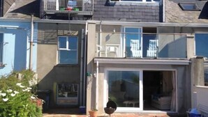 Une maison à deux étages dotée d’un balcon, d’une terrasse agrémentée de plantes en pot et d’une porte-fenêtre coulissante donnant sur un espace extérieur abrité.
