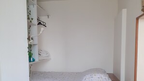 Habitació petita i senzilla amb un llit individual, una taula de nit, una prestatgeria amb roba penjada i un petit tamboret.