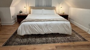 A bedroom with a large bed, two bedside tables, a nightstand, a window with curtains, and a wooden floor. - Steel City Stay- 4B/2B Getaway (Pittsburgh)