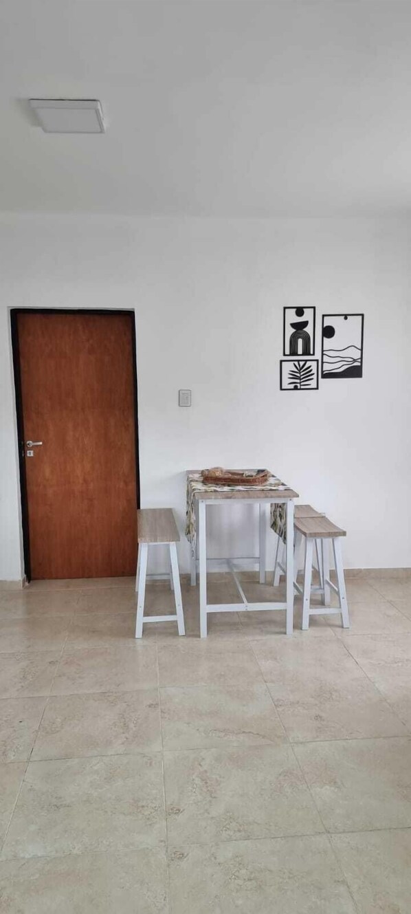 A room with a wooden door, a small table with a tablecloth, two white stools, and three framed pictures on the wall. - Center Cruz del Eje (Cruz del Eje)