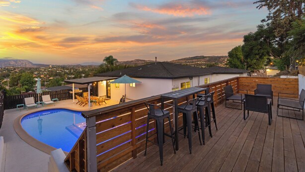 Zona de piscina con terraza de madera, taburetes de bar y un área de estar cubierta con sombrillas.