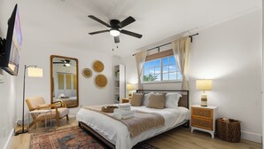 Un dormitorio con cama, ventilador de techo, televisor, silla, mesita de noche con lámpara y ventana con cortinas.