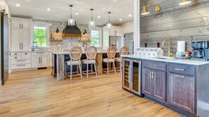 Een moderne keuken met houten vloeren, witte kasten, een centraal eiland met stoelen, hanglampen en een accentmuur van baksteen.