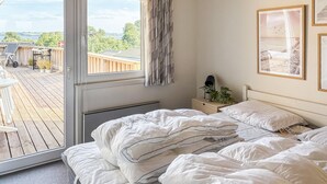 Une chambre avec une grande fenêtre, un lit avec une literie blanche, une table de chevet avec une lampe, et une vue sur une terrasse en bois et des meubles d’extérieur.