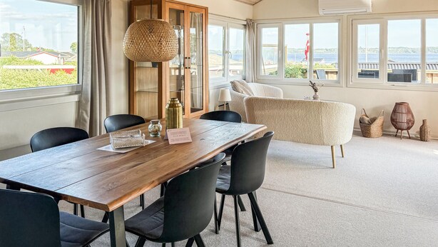 Un espace repas avec une table en bois, des chaises noires, un luminaire suspendu en fibres tressées et une vue sur la mer grâce à de grandes fenêtres.