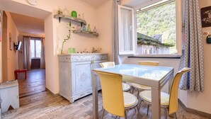 Une cuisine avec une table et des chaises, une fenêtre donnant sur un espace verdoyant et une étagère contenant des ustensiles de cuisine.