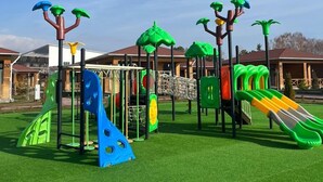 A playground with various play structures including slides and climbing areas, surrounded by a grassy field and buildings in the background. - Baytur Resort & Spa (Cholpon-Ata)
