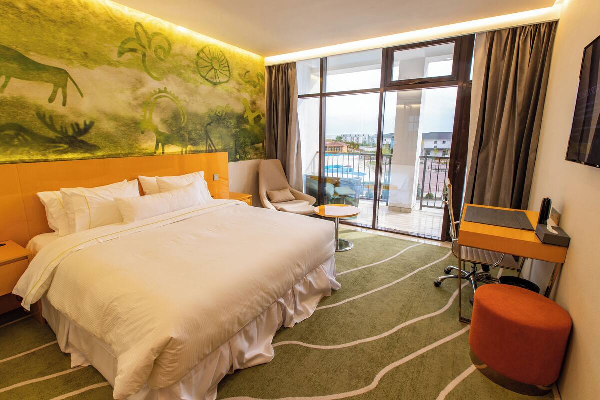 A hotel room with a large bed, a desk with a laptop, a chair, and a view of a pool through a glass door.