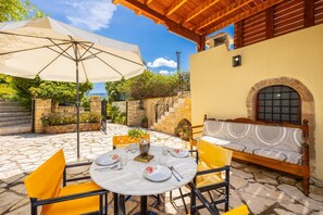 An outdoor patio with a round table set for dining, yellow chairs, a cushioned bench, and a large umbrella.