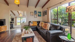 A modern living room with a leather sofa, a coffee table, and a TV. There are framed pictures on the wall and a large window with a view of greenery. - Cozy Lake Retreat - Games Room, Spa, Sleeps 6 (Hamurana)