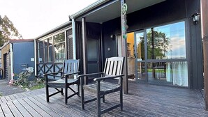 A wooden deck with two chairs, a covered porch, and a large glass door leading to an interior space. - Cozy Lake Retreat - Games Room, Spa, Sleeps 6 (Hamurana)