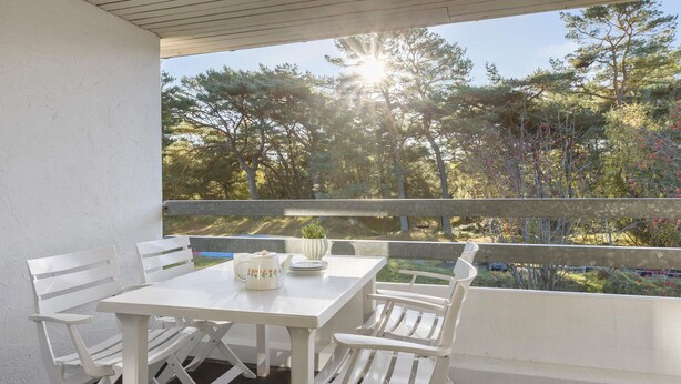 A balcony with a white table and chairs, a potted plant, and a view of trees and a lake. - Lovely apartment in Nexø with WiFi (Nexø)