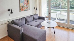 A modern living room with a grey sectional sofa, a round white coffee table, and a large window with a view of trees. - Lovely apartment in Nexø with WiFi (Nexø)
