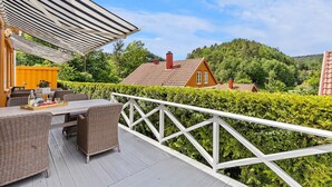 A wooden deck with outdoor furniture, a white railing, and a view of a forested area with houses.